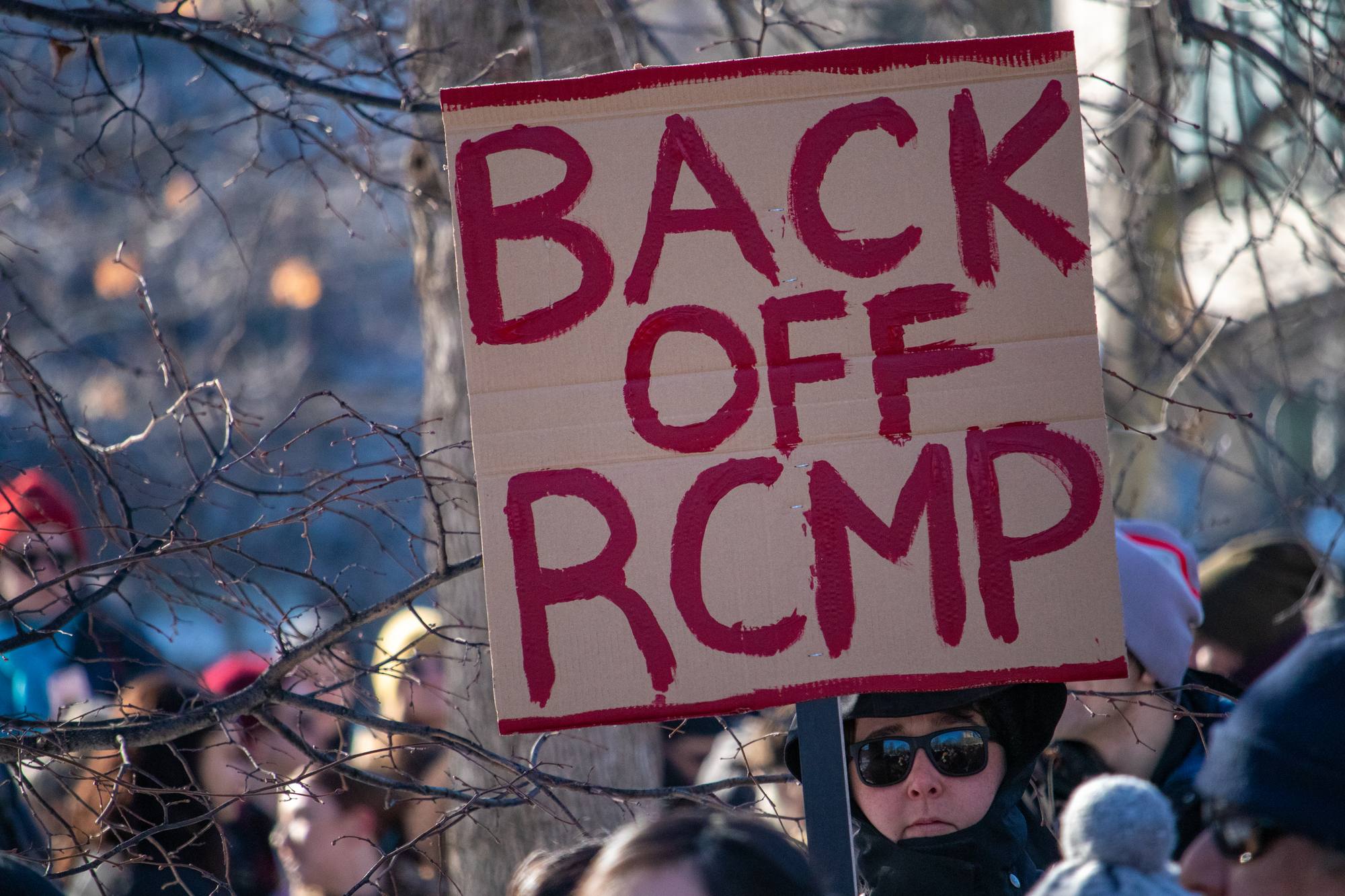 Person holding sign that reads Back off RCMP regarding the RCMP discrimination class action lawsuit expanding