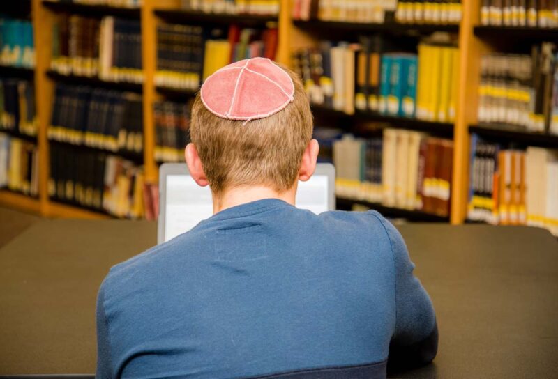 Back view of a male wearing a yarmulke studying on his laptop, representing the anti-semitic universities class action.
