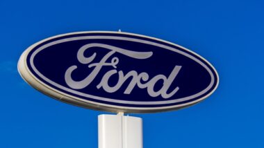 The iconic blue oval logo of Ford Motor Company is seen on a tall dealership sign against a clear blue sky