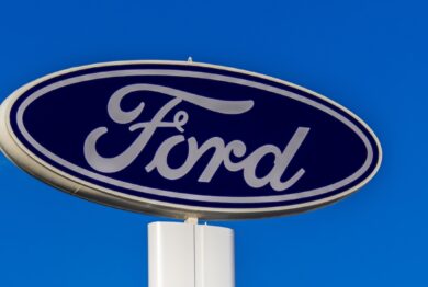 The iconic blue oval logo of Ford Motor Company is seen on a tall dealership sign against a clear blue sky