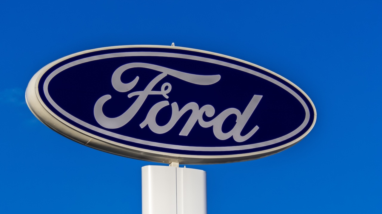 The iconic blue oval logo of Ford Motor Company is seen on a tall dealership sign against a clear blue sky